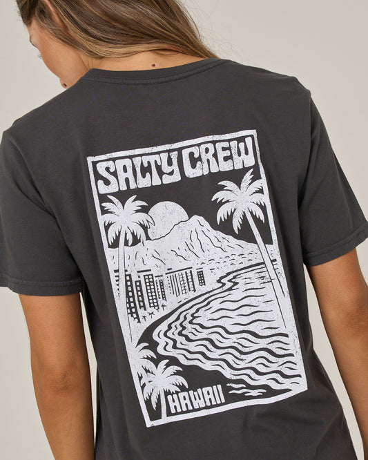 A woman with long hair wears the Salty Crew Aloha Breeze Boyfriend Tee in charcoal, a relaxed fit short sleeve style crafted from soft cotton jersey. The back features a white graphic of palm trees, mountains, water, and buildings along with the words “SALTY CREW” and “HAWAII.”.