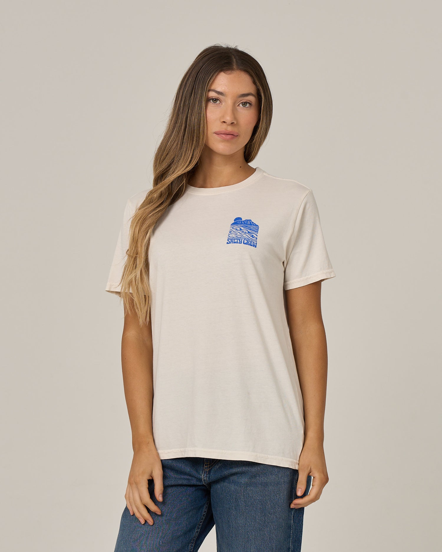 A woman with long, wavy hair wears the Salty Crew Aloha Breeze Boyfriend Tee in Off White—a relaxed fit tee crafted from soft cotton jersey with a small blue chest graphic—styled with blue jeans and standing against a plain light background.