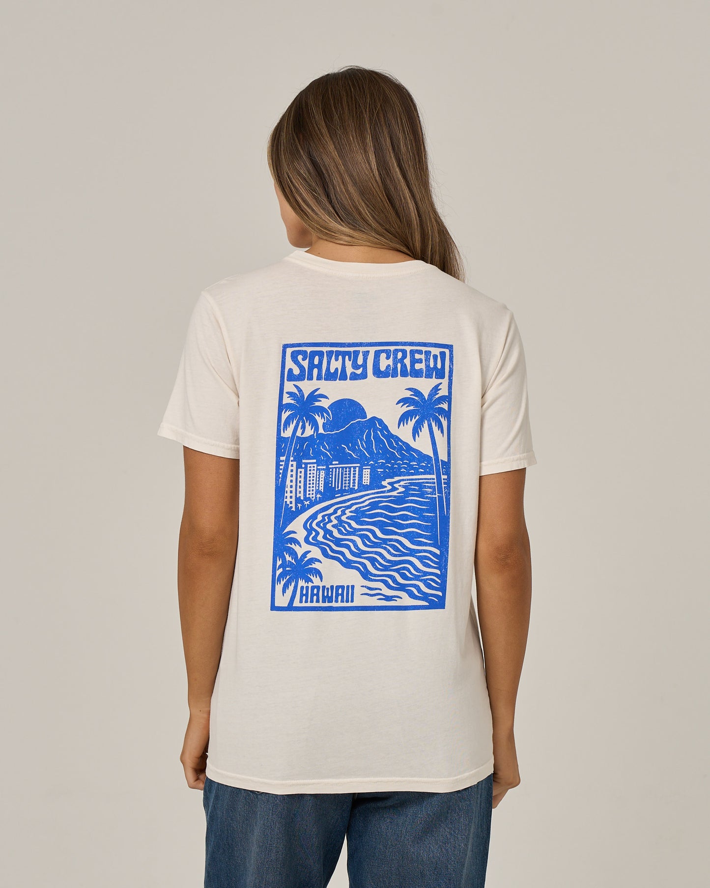 A person wearing the Salty Crew Aloha Breeze Boyfriend Tee in Off White—a relaxed fit short sleeve T-shirt made from soft cotton jersey. The tee features a white base with a blue ocean scene graphic, palm trees, and “Salty Crew Hawaii” printed on the back. The shirt is showcased against a plain background.