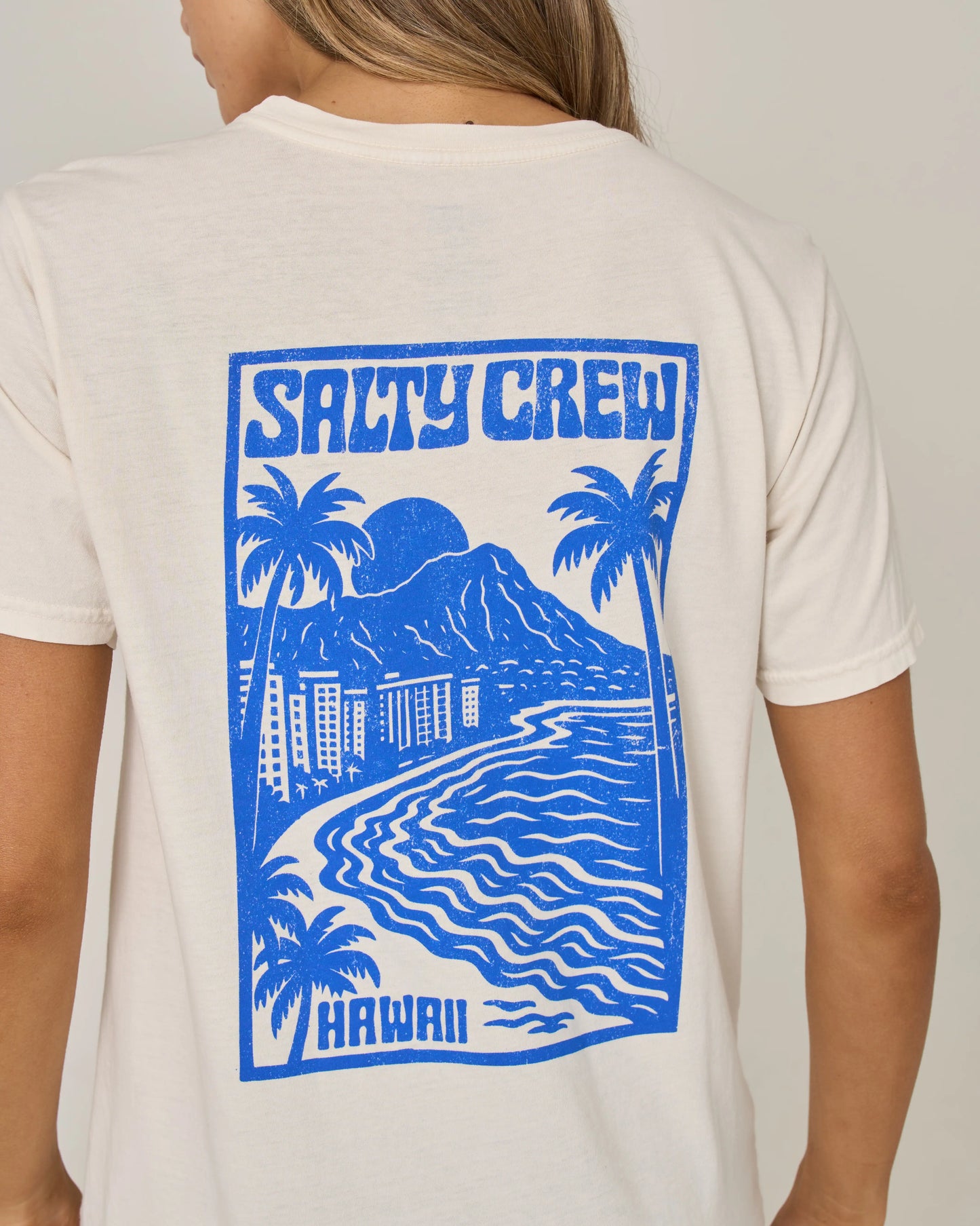 A person wearing the Salty Crew Aloha Breeze Boyfriend Tee in off white—a relaxed fit short sleeve tee crafted from soft cotton jersey, featuring a blue graphic of palm trees, ocean waves, mountains, and buildings on the back with “Salty Crew” and “Hawaii” text.