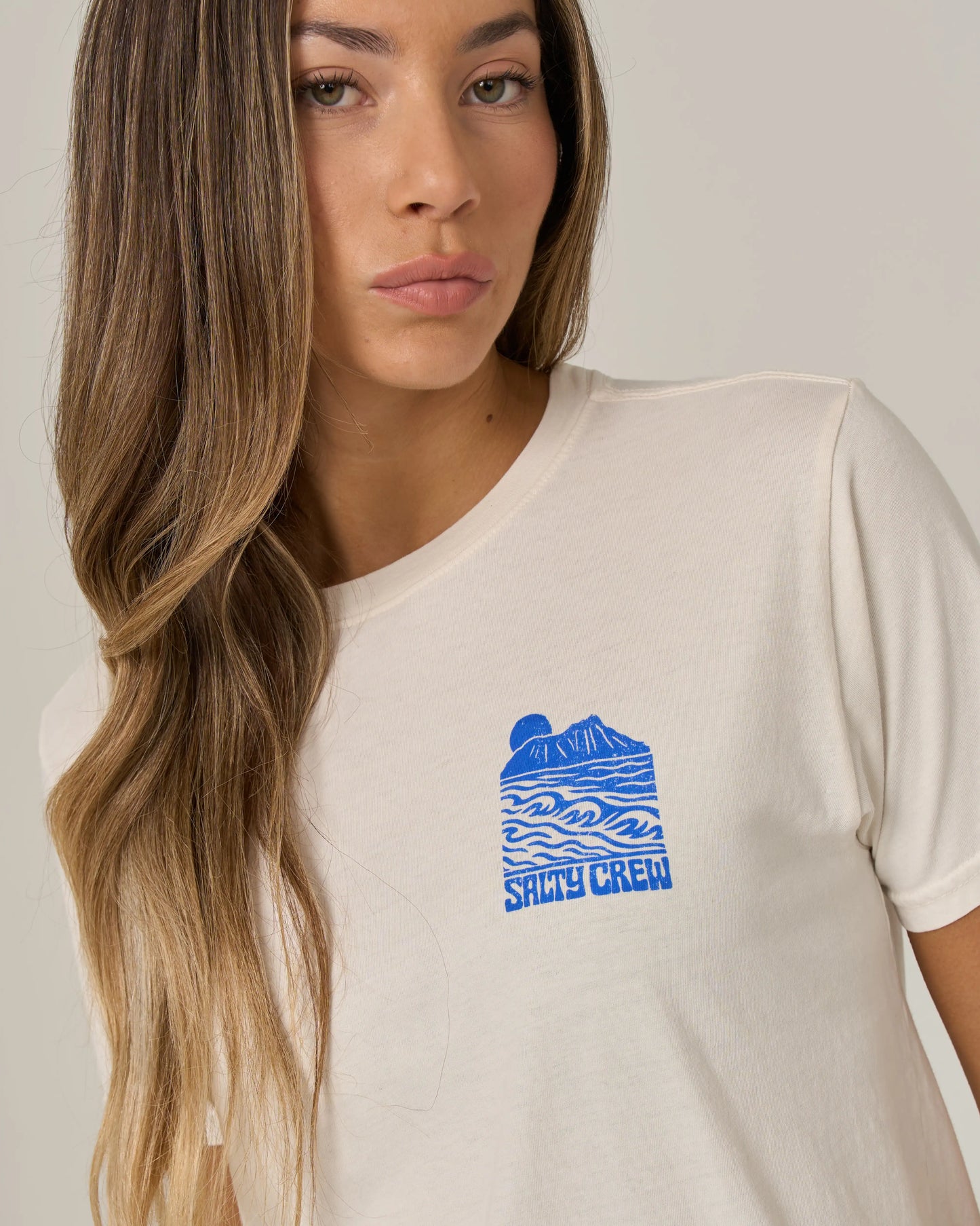 A woman with long brown hair wears the Salty Crew Aloha Breeze Boyfriend Tee in Off White. This relaxed fit, short sleeve t-shirt is crafted from soft cotton jersey and features a blue wave and mountain graphic on the left chest. She stands against a plain light background, looking slightly to the side, embodying casual comfort and style.