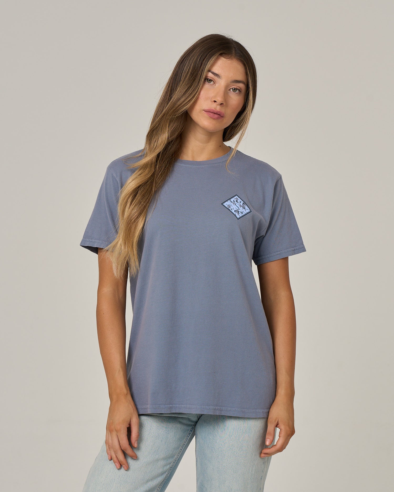 A woman with long, straight hair stands against a plain, light background wearing the Salty Crew Tippet Fill Boyfriend Tee in Flint. Featuring a small graphic on the left chest, this blue cotton jersey shirt has short sleeves and a relaxed fit that adds to her comfortable, effortless look. Her expression is neutral and her arms are relaxed by her sides.