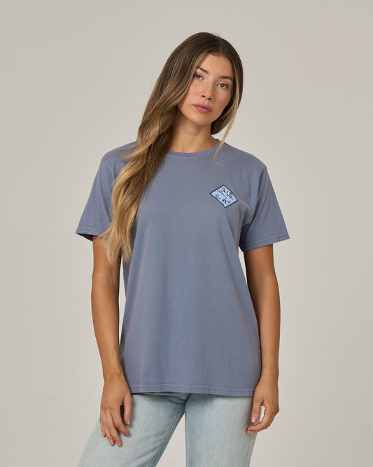 A woman with long, straight hair stands against a plain, light background wearing the Salty Crew Tippet Fill Boyfriend Tee in Flint. Featuring a small graphic on the left chest, this blue cotton jersey shirt has short sleeves and a relaxed fit that adds to her comfortable, effortless look. Her expression is neutral and her arms are relaxed by her sides.
