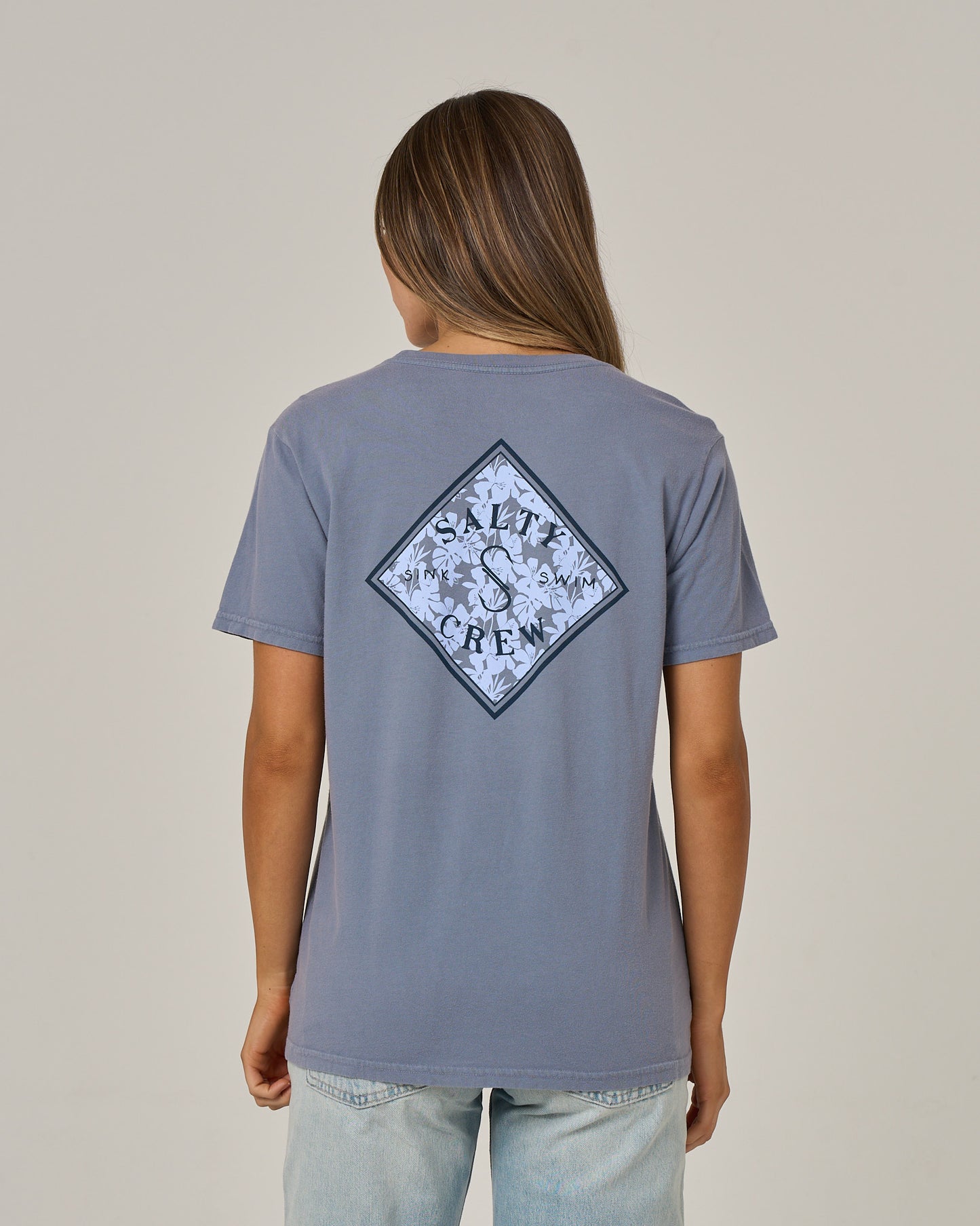 A person with long brown hair is wearing the Salty Crew Tippet Fill Boyfriend Tee in Flint—a blue, short-sleeve cotton jersey T-shirt featuring a diamond-shaped Salty Crew logo with a floral design on the back—standing against a plain background.