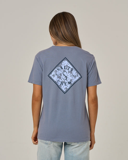 A person with long brown hair is wearing the Salty Crew Tippet Fill Boyfriend Tee in Flint—a blue, short-sleeve cotton jersey T-shirt featuring a diamond-shaped Salty Crew logo with a floral design on the back—standing against a plain background.