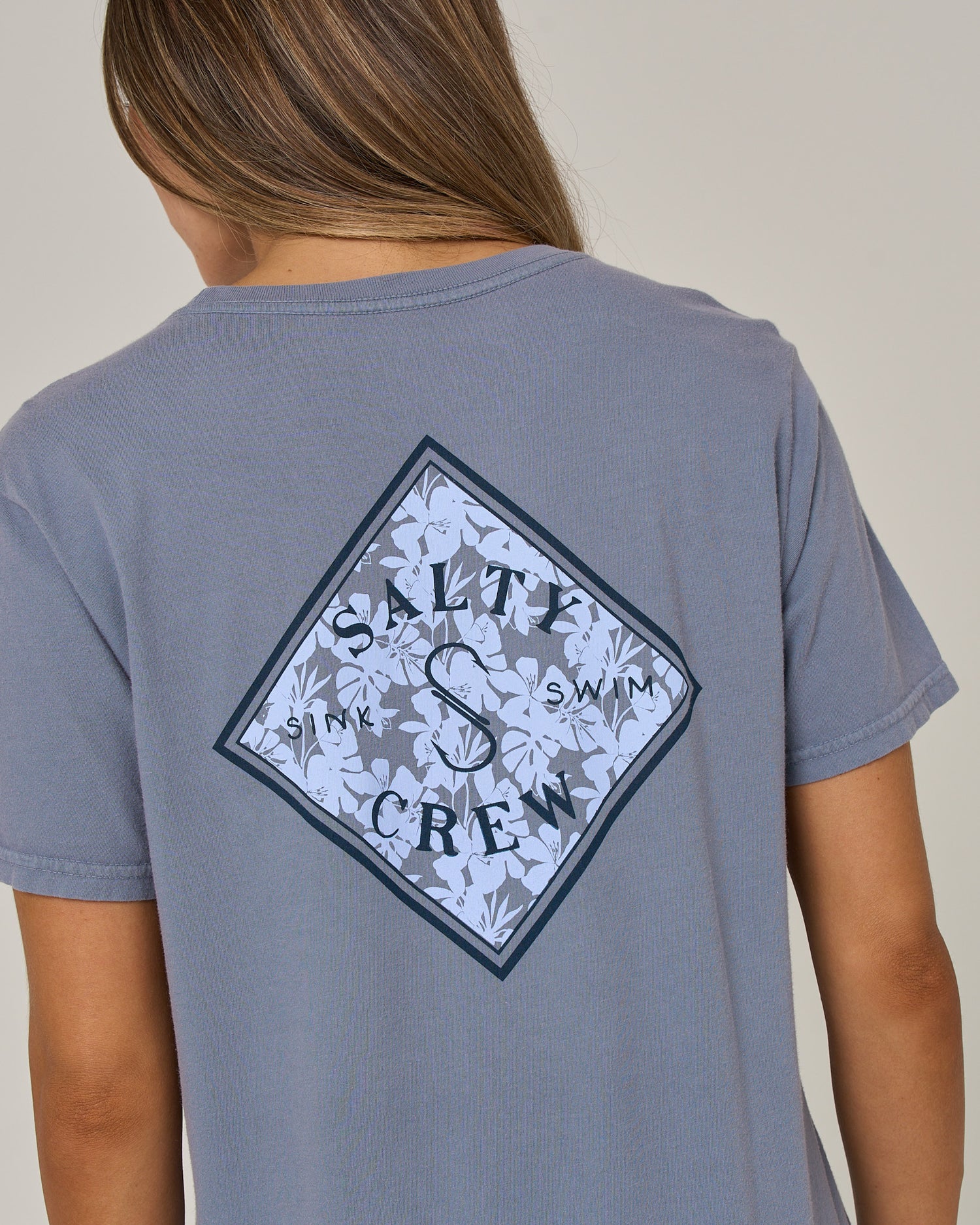 A person is seen from behind, wearing the Salty Crew Tippet Fill Boyfriend Tee in Flint—a relaxed fit gray cotton jersey shirt featuring a diamond-shaped floral graphic and bold SALTY CREW lettering across the back. The short sleeves reveal only their back and part of their brown hair as they turn away.