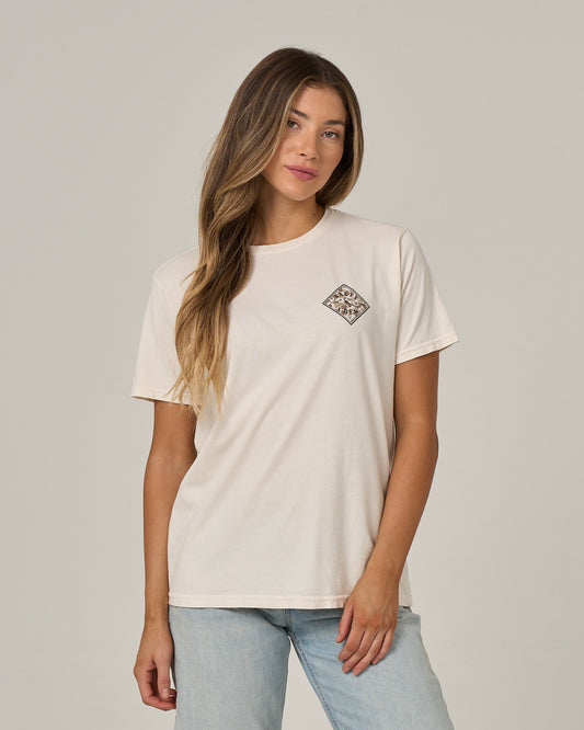 A woman with long, wavy hair models the Salty Crew Tippet Fill Boyfriend Tee in Off White—a relaxed fit, cotton jersey T-shirt featuring short sleeves and a small patterned patch on the left chest. She pairs it with light blue jeans and stands against a simple, light background, effortlessly showcasing the laid-back style of this tee.