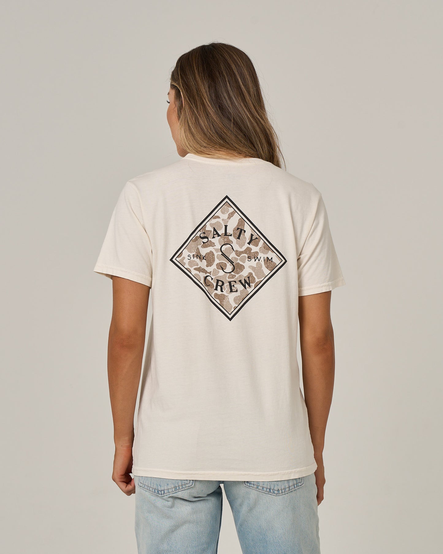 A person with long hair wears a Salty Crew Tippet Fill Boyfriend Tee in Off White, featuring a relaxed fit and short sleeves, paired with light blue jeans. They are facing away to display the back logo, standing against a plain light background.