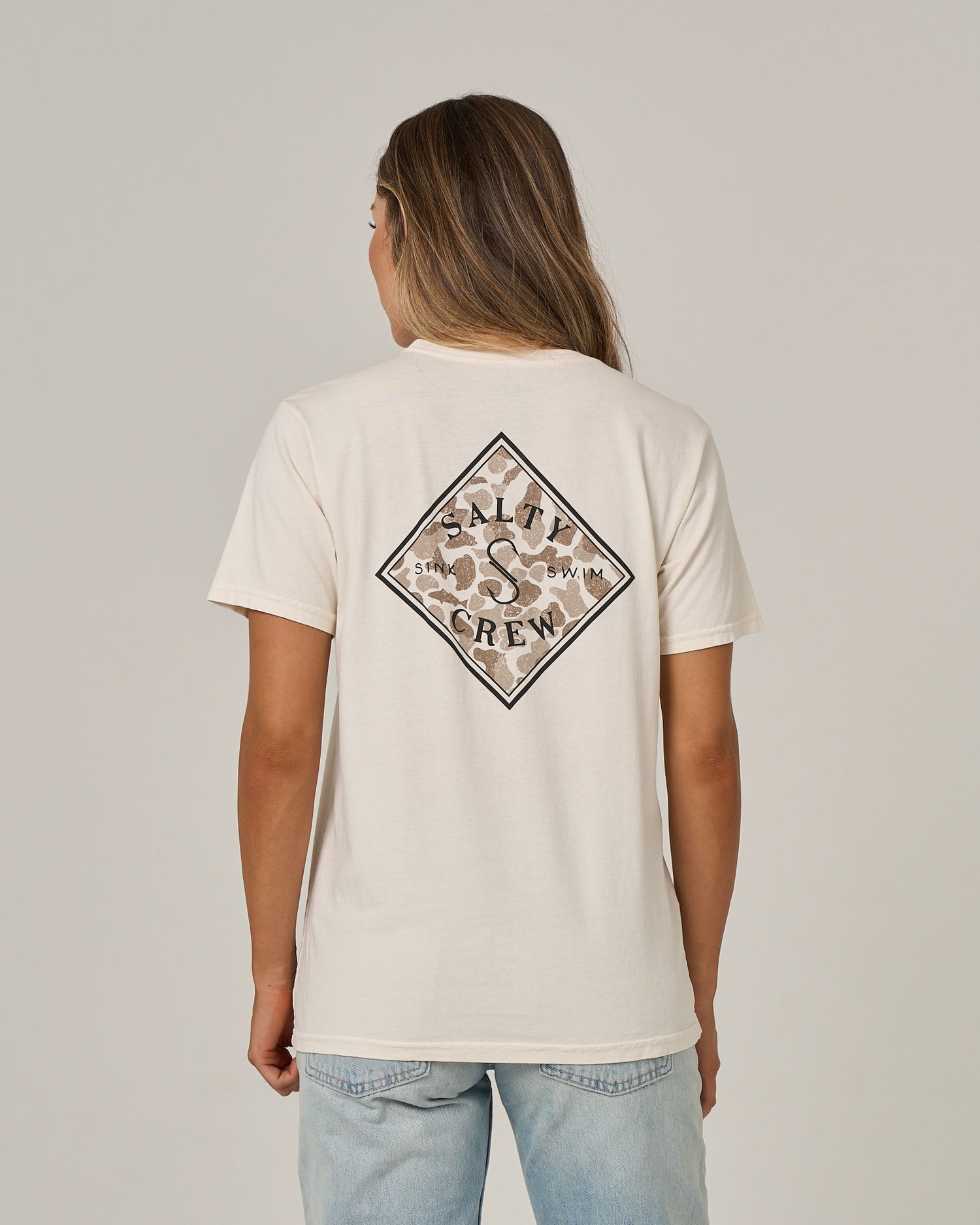 A person with long hair wears a Salty Crew Tippet Fill Boyfriend Tee in Off White, featuring a relaxed fit and short sleeves, paired with light blue jeans. They are facing away to display the back logo, standing against a plain light background.