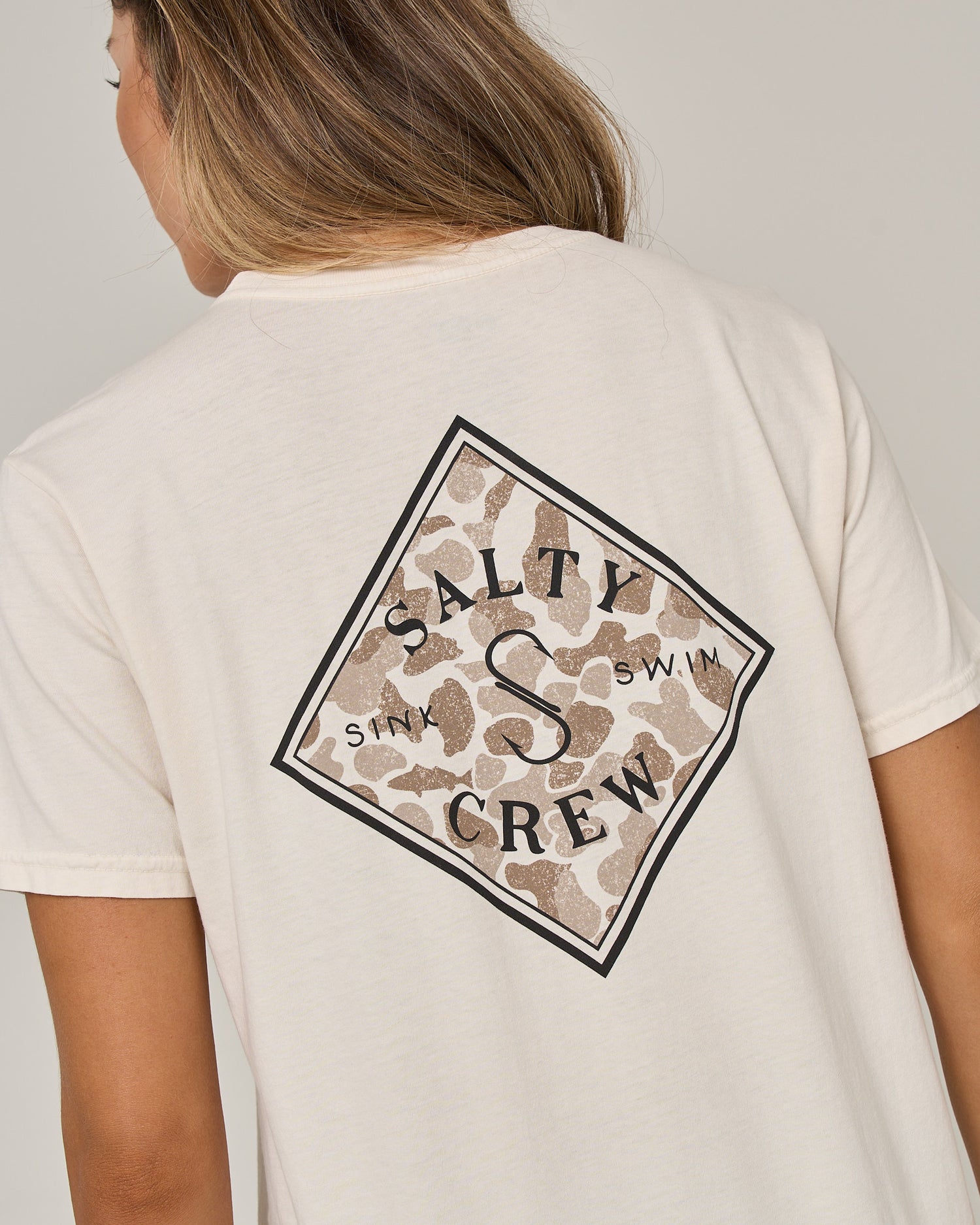 A person is shown from the back wearing the Salty Crew Tippet Fill Boyfriend Tee in Off White, a relaxed fit, short sleeve cotton jersey featuring a diamond-shaped Salty Crew logo on the back with a tan and brown camouflage background and the words “Sink and Swim.”.