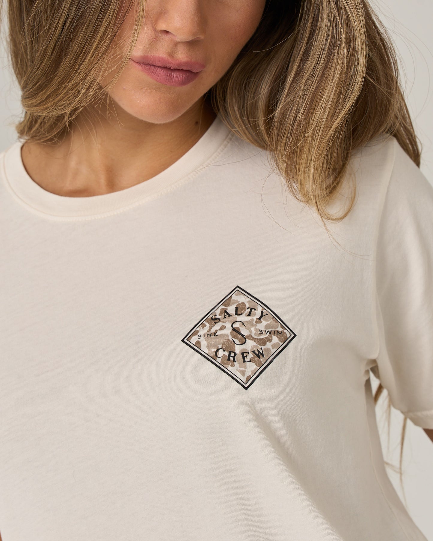 A woman with long, light brown hair wears the Salty Crew Tippet Fill Boyfriend Tee in Off White, a relaxed fit, short sleeve cotton jersey shirt featuring a small diamond-shaped camouflage Salty Crew logo on the chest. This tee combines comfort and effortless style.