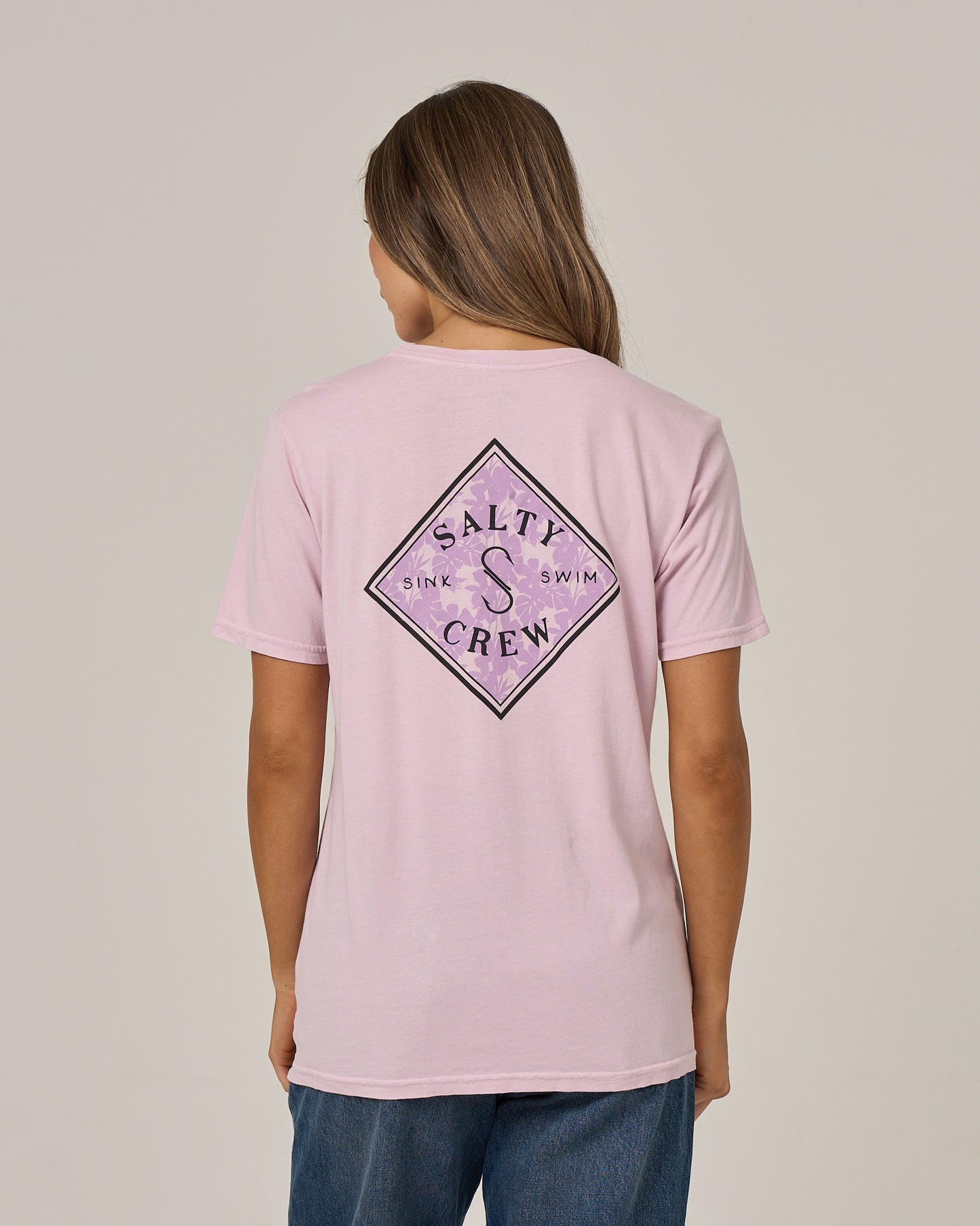 A person with long hair is seen from behind wearing the Salty Crew Tippet Fill Boyfriend Tee in Pale Orchid—a relaxed-fit, light pink t-shirt featuring a diamond-shaped logo and “Sink or Swim” screenprint on the back—paired with blue jeans.