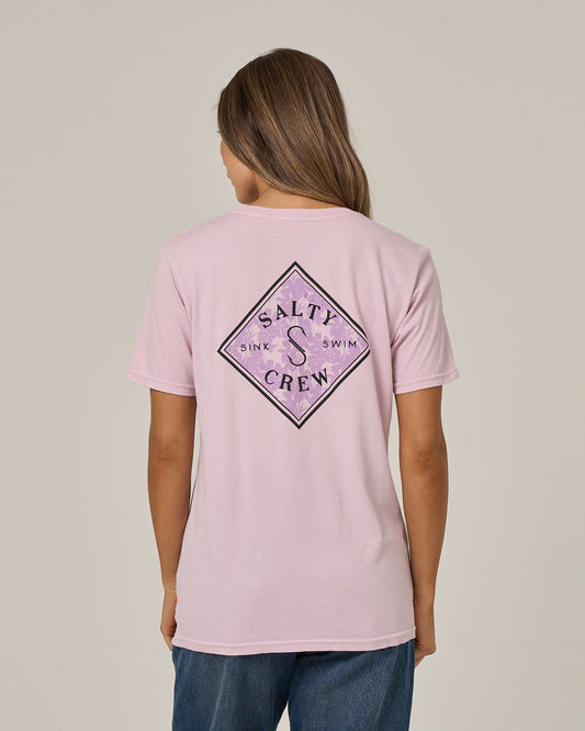 A person with long hair is seen from behind wearing the Salty Crew Tippet Fill Boyfriend Tee in Pale Orchid—a relaxed-fit, light pink t-shirt featuring a diamond-shaped logo and “Sink or Swim” screenprint on the back—paired with blue jeans.