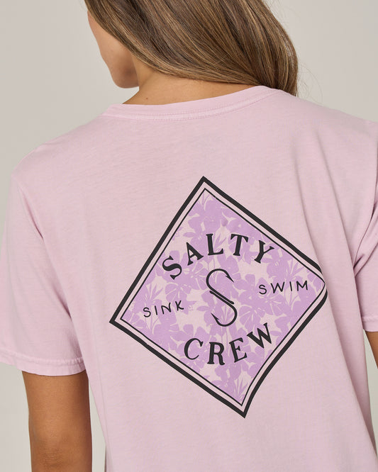 The back of a person wearing the Salty Crew Tippet Fill Boyfriend Tee in Pale Orchid—a relaxed fit, light pink t-shirt featuring a diamond-shaped floral logo with “Salty Crew Sink Swim” in black text. Crafted from soft cotton jersey, this screen-printed tee pairs effortlessly with the person’s loose brown hair for an easygoing look.