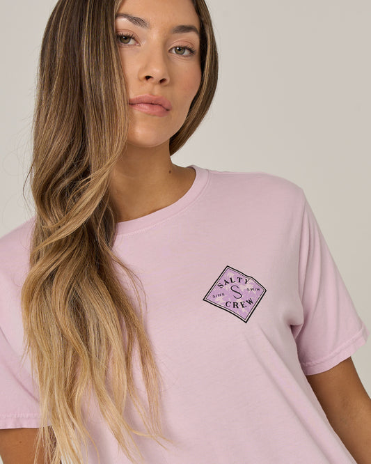 A woman with long, wavy blonde hair is wearing the Salty Crew Tippet Fill Boyfriend Tee in Pale Orchid, standing against a plain light background and looking at the camera with a neutral expression.