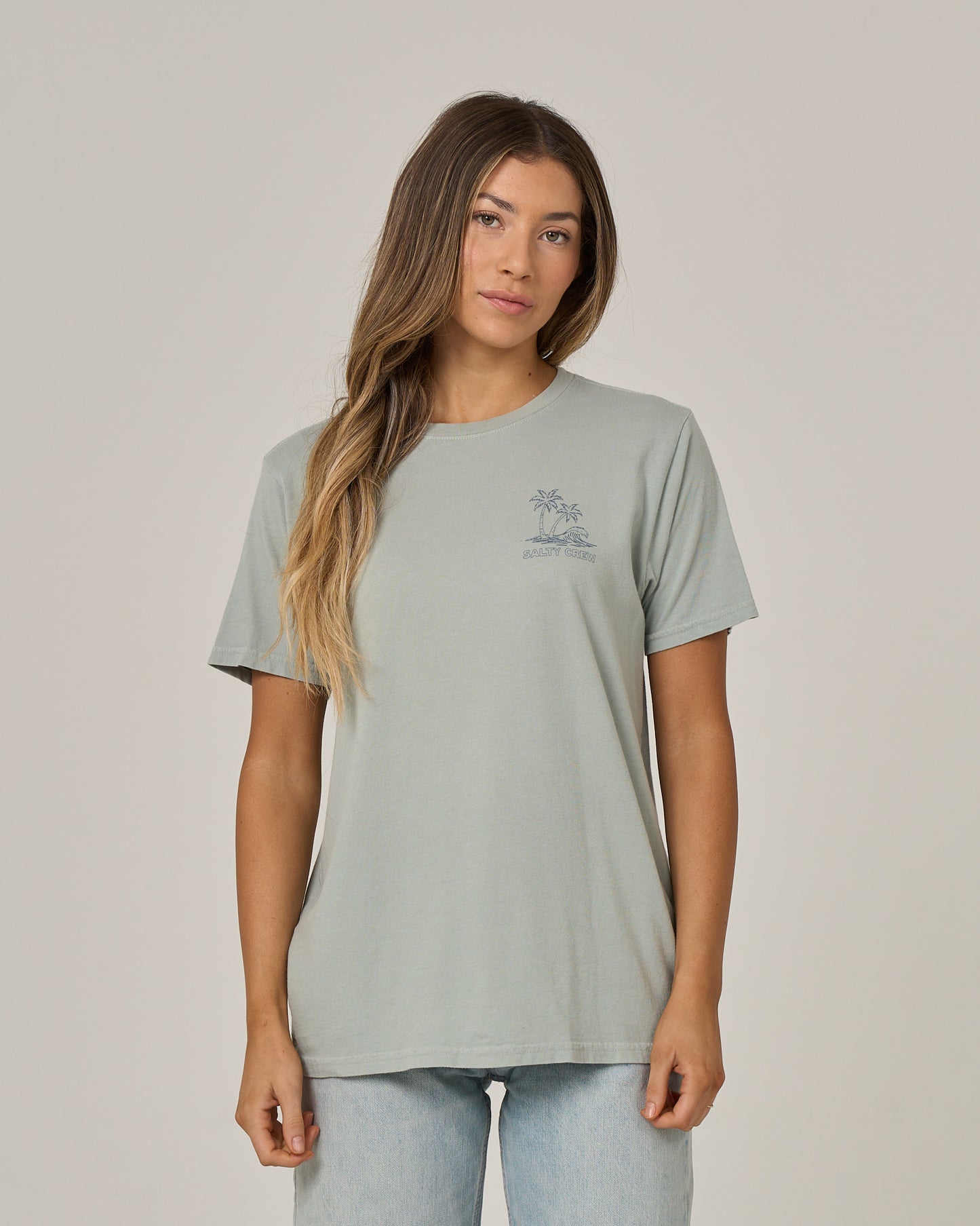 A woman with long, wavy hair is wearing the Salty Crew Island Wave Boyfriend Tee in Aqua Grey—a soft cotton jersey short sleeve t-shirt—paired with light blue jeans. She stands against a plain background facing forward with a neutral expression.