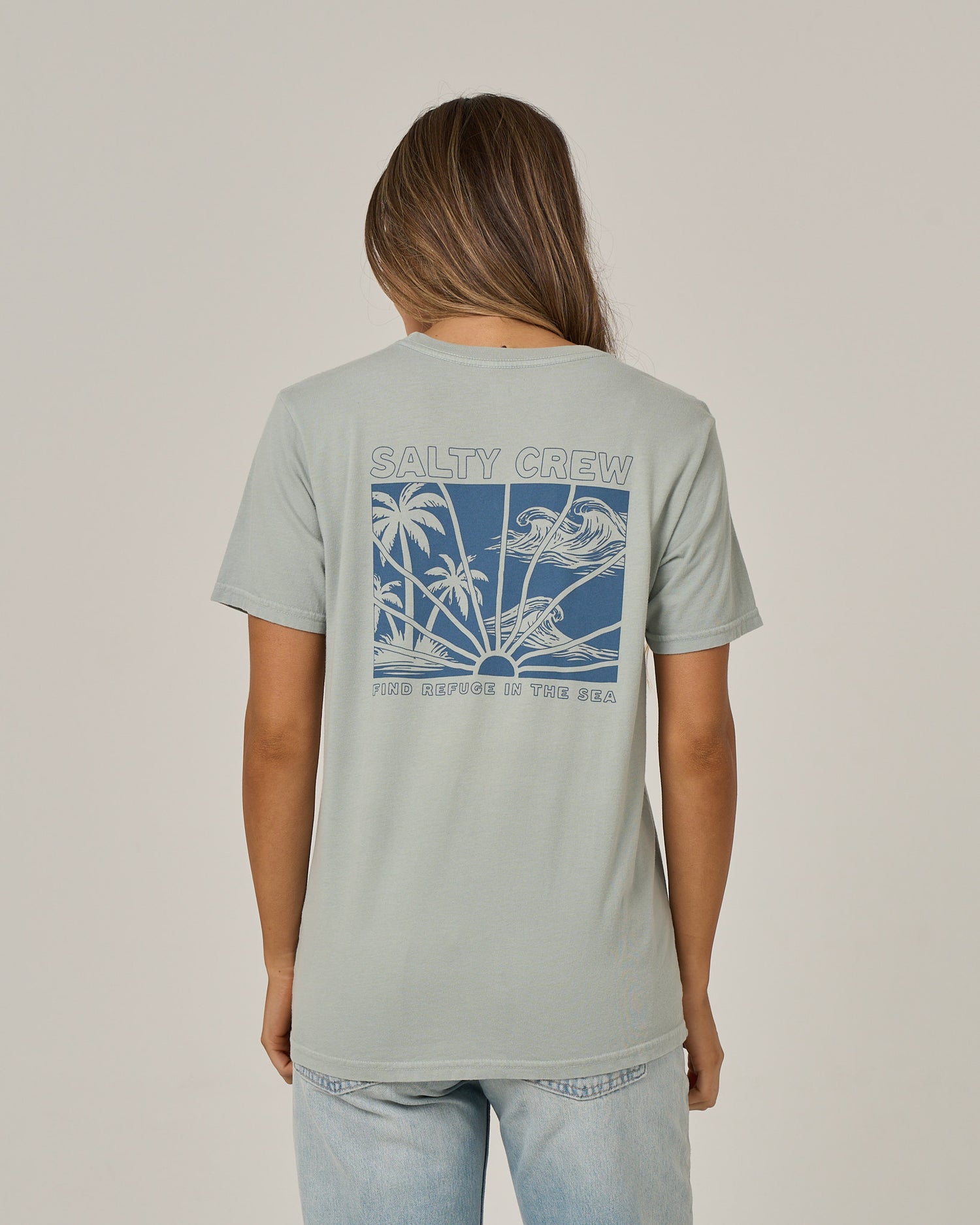 A person with long hair, dressed in jeans and a Salty Crew Island Wave Boyfriend Tee in Aqua Grey, is shown from the back. The relaxed fit short sleeve t-shirt, made from soft cotton jersey, features a blue graphic with palm trees, a bird, and the phrase “Find refuge in the sea.”.