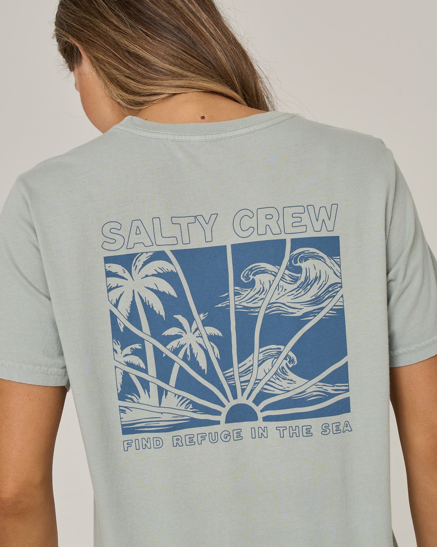 A person wearing the Salty Crew Island Wave Boyfriend Tee in Aqua Grey, a light blue cotton jersey relaxed fit t-shirt featuring a graphic of waves, palm trees, and a sunset with the text “SALTY CREW” and “FIND REFUGE IN THE SEA.”.