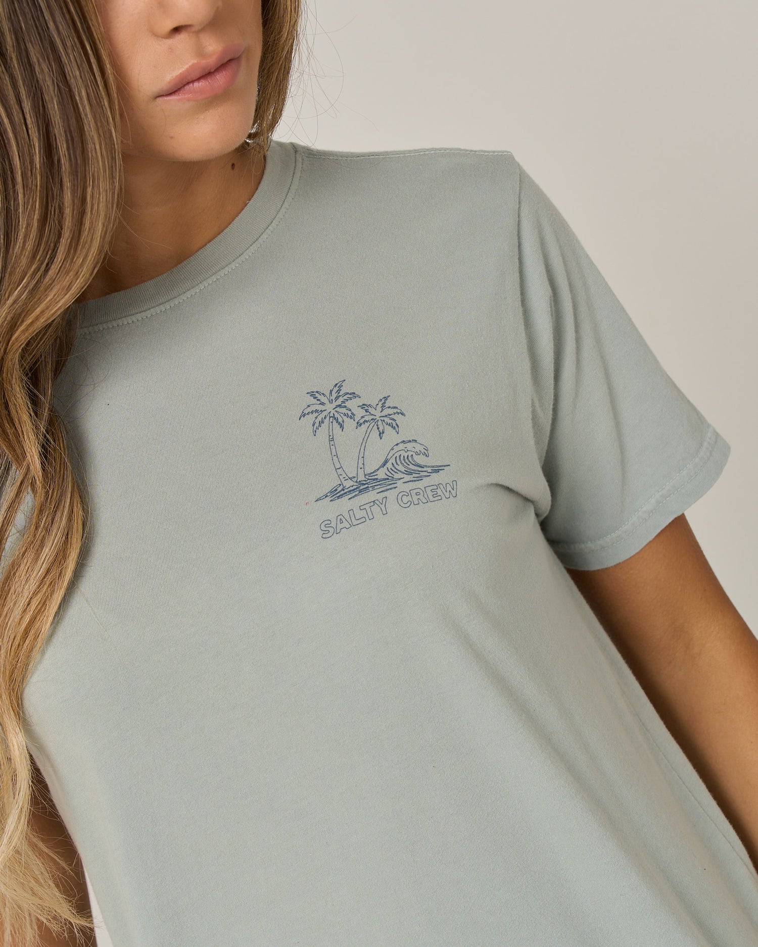 A woman is wearing the Salty Crew Island Wave Boyfriend Tee in Aqua Grey—a relaxed fit top crafted from soft cotton jersey, featuring a small palm tree and wave graphic above the SALTY CREW logo on the chest. Only her head and upper torso are visible.