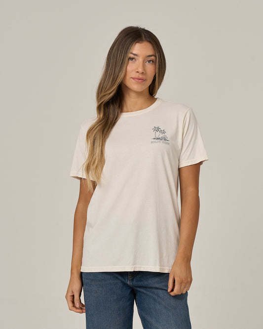 A woman with long, wavy hair wears the Salty Crew Island Wave Boyfriend Tee in off white—a relaxed fit tee crafted from soft cotton jersey with a small chest graphic—paired with blue jeans, standing against a plain light background.