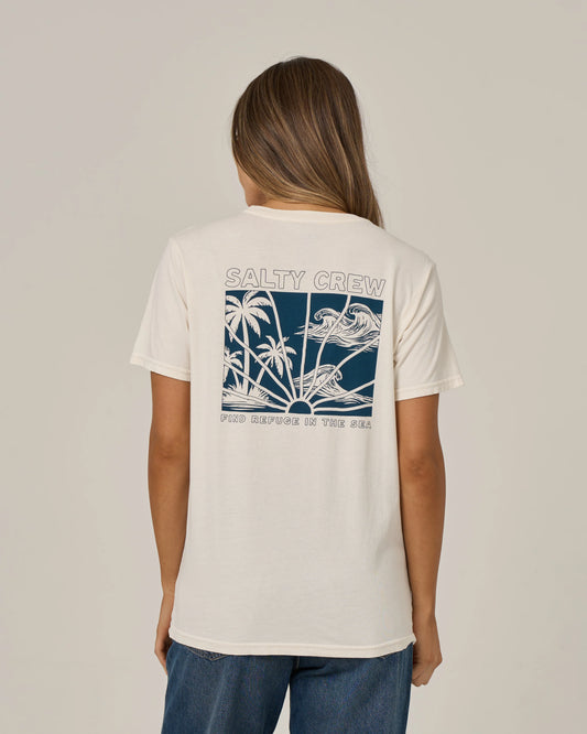 A woman with long hair is wearing the Salty Crew Island Wave Boyfriend Tee in Off White—a relaxed fit, cotton jersey shirt featuring a blue and white tropical graphic with text on the back. She pairs it with blue jeans and is standing facing away to highlight the tee’s back design.