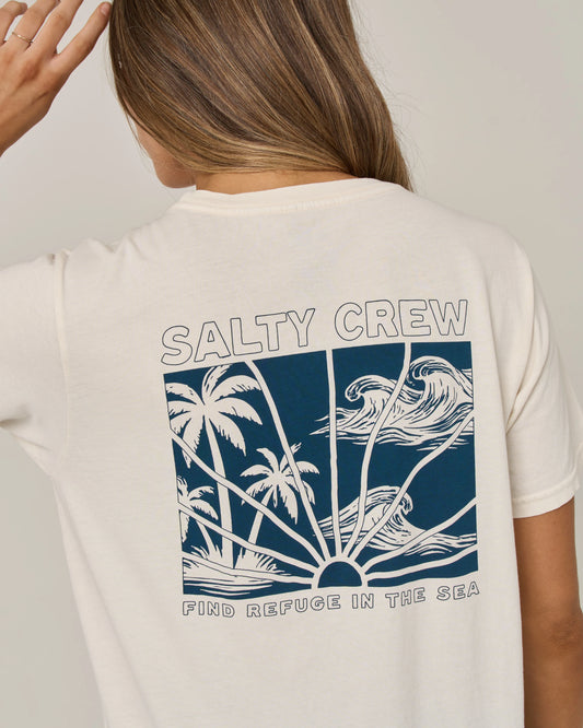 A person wearing the Salty Crew Island Wave Boyfriend Tee in off white—a relaxed fit short sleeve shirt crafted from soft cotton jersey, featuring a blue graphic of waves, palm trees, and a sunset along with the text “SALTY CREW” and “FIND REFUGE IN THE SEA.”.