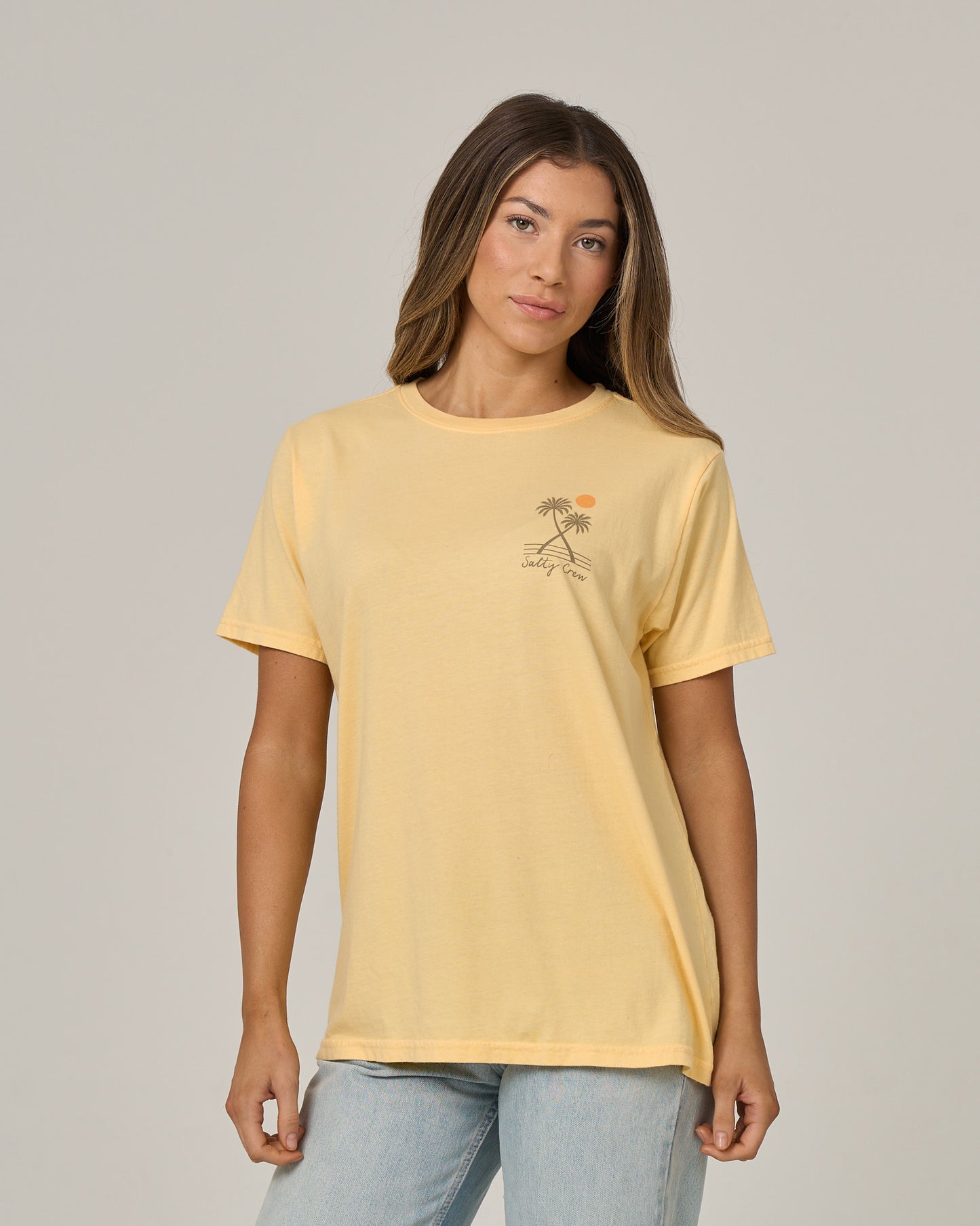 A woman wearing the Salty Crew Moon Wake Boyfriend Tee in Custard, featuring a small palm tree and sun graphic on the left chest, stands against a plain light background.