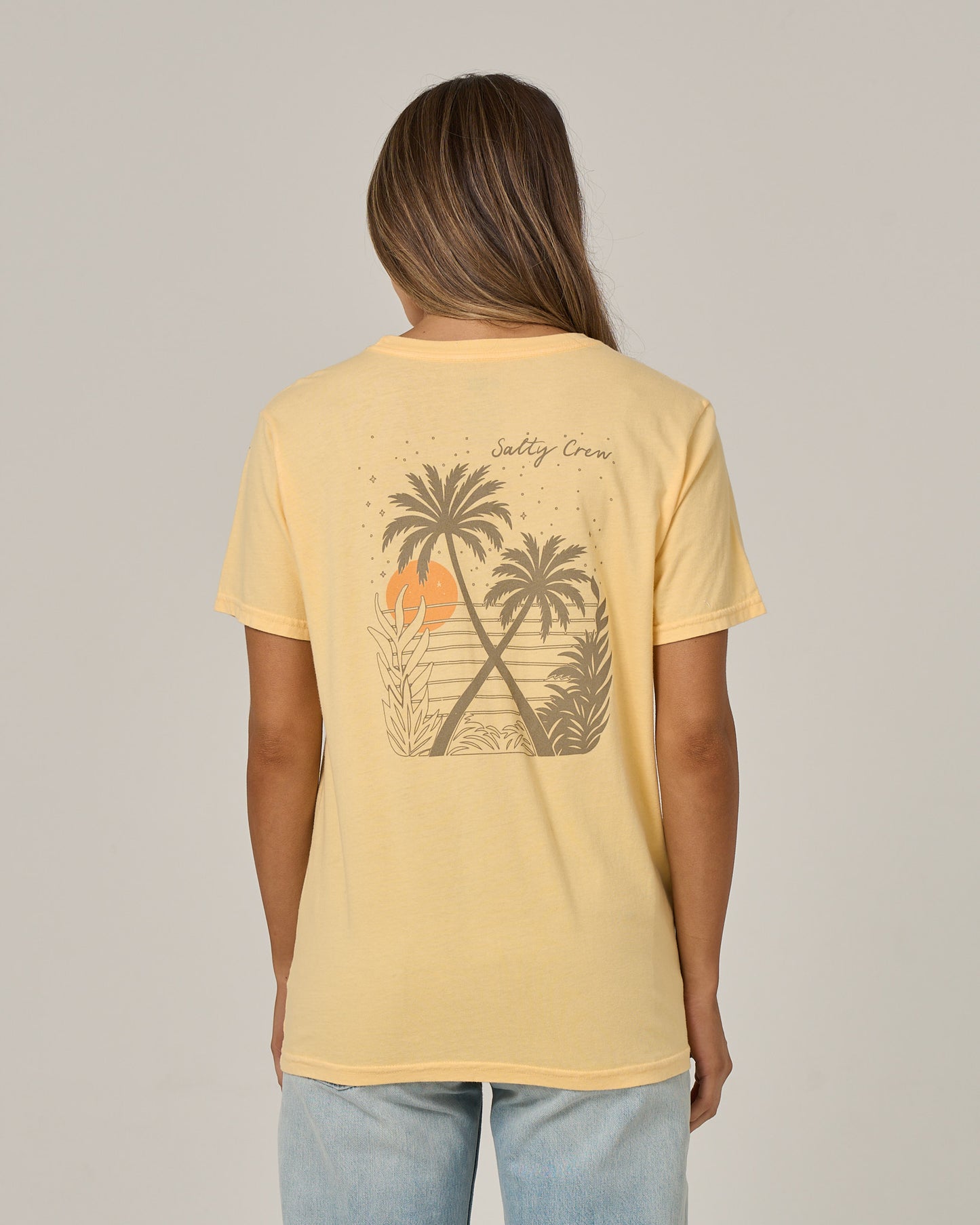 A woman with long hair, facing away from the camera, wears the Salty Crew Moon Wake Boyfriend Tee in Custard—a relaxed-fit, short sleeve cotton jersey shirt featuring a graphic of palm trees, sunset, and waves on the back. She pairs it with light blue jeans.