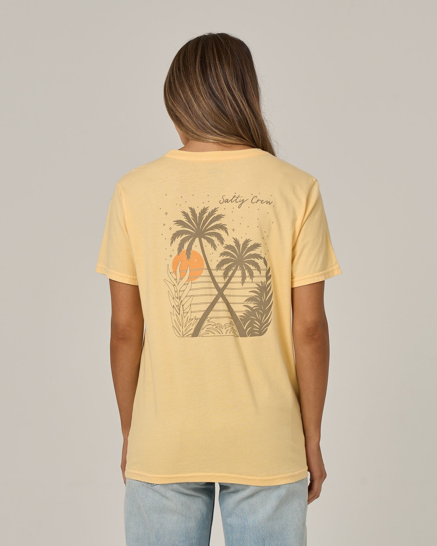 A woman with long hair, facing away from the camera, wears the Salty Crew Moon Wake Boyfriend Tee in Custard—a relaxed-fit, short sleeve cotton jersey shirt featuring a graphic of palm trees, sunset, and waves on the back. She pairs it with light blue jeans.