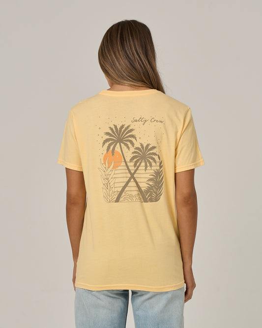 A woman with long hair, facing away from the camera, wears the Salty Crew Moon Wake Boyfriend Tee in Custard—a relaxed-fit, short sleeve cotton jersey shirt featuring a graphic of palm trees, sunset, and waves on the back. She pairs it with light blue jeans.