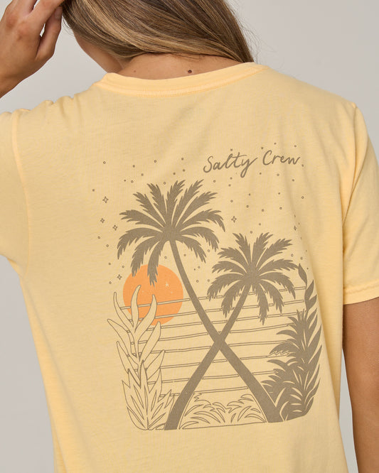 A person is seen from the back wearing the Salty Crew Moon Wake Boyfriend Tee in Custard, a comfortable cotton jersey shirt with a relaxed fit and short sleeves. The back features a graphic of two crossed palm trees, a sunset, and tropical leaves surrounded by small star icons, with the words “Salty Crew” in cursive above the design.