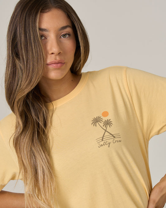 A young woman with long, wavy hair is wearing the Salty Crew Moon Wake Boyfriend Tee in Custard—a relaxed fit, soft cotton jersey shirt featuring a palm tree and sun graphic along with the Salty Crew logo.