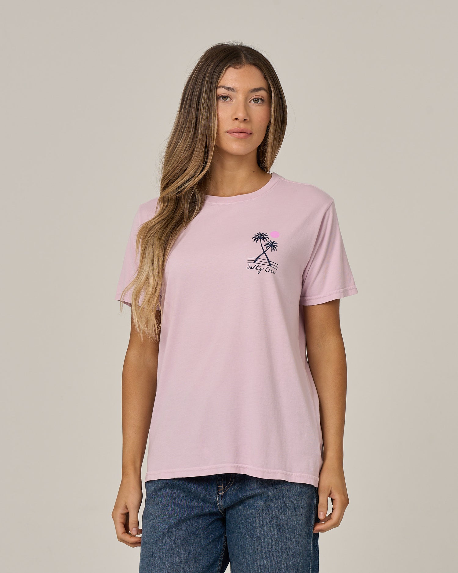 A woman with long hair wears the Salty Crew Moon Wake Boyfriend Tee in Pale Orchid—a light pink cotton jersey relaxed-fit t-shirt featuring a small palm tree graphic and text on the chest—paired with blue jeans and standing against a plain beige background.