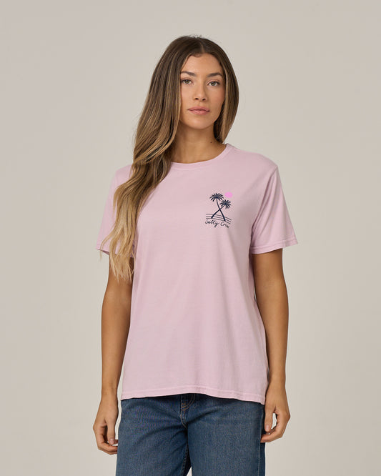 A woman with long hair wears the Salty Crew Moon Wake Boyfriend Tee in Pale Orchid—a light pink cotton jersey relaxed-fit t-shirt featuring a small palm tree graphic and text on the chest—paired with blue jeans and standing against a plain beige background.