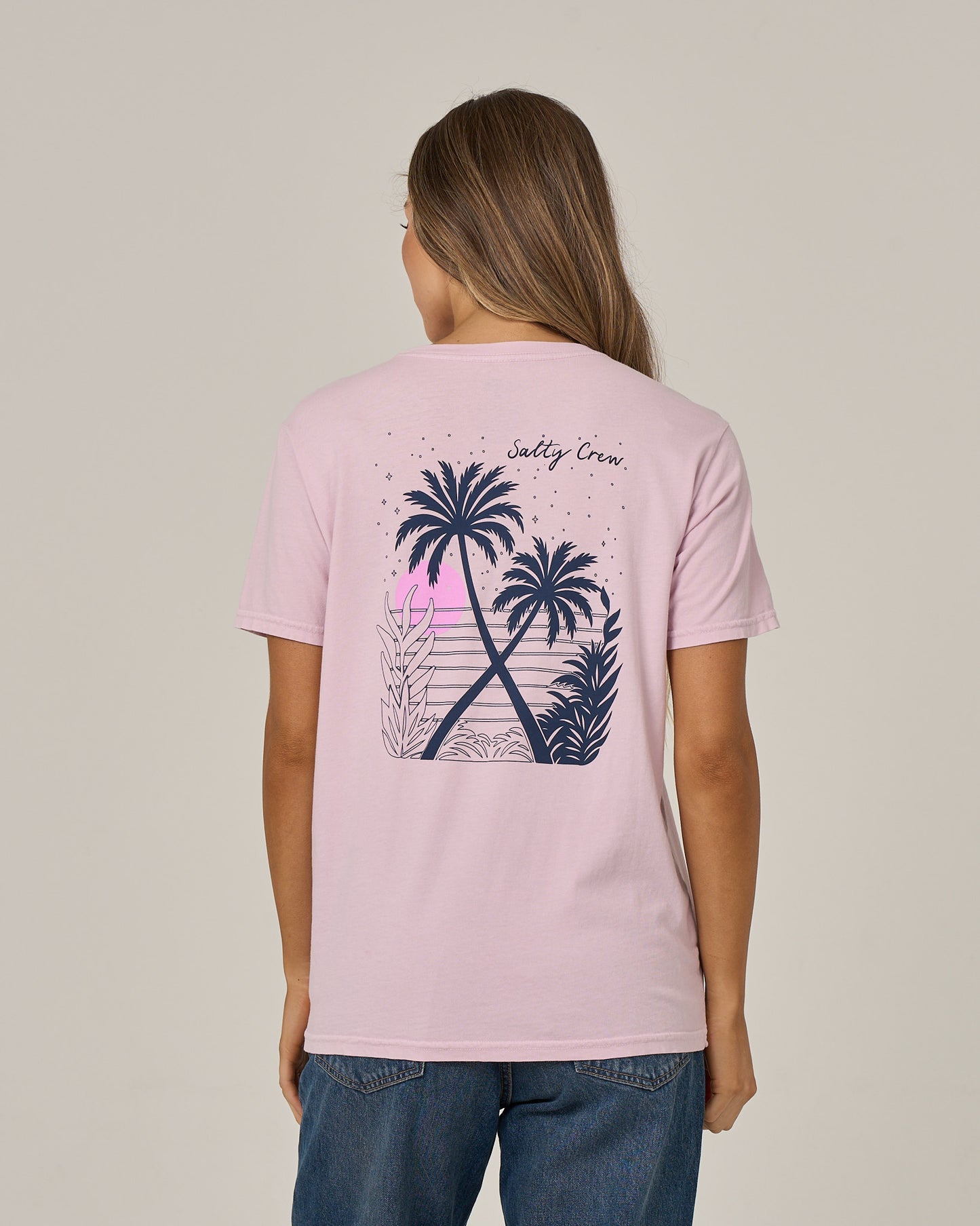 A woman with long hair, dressed in blue jeans and facing away from the camera, showcases the relaxed fit of the Salty Crew Moon Wake Boyfriend Tee in Pale Orchid—a light pink, short sleeve cotton jersey T-shirt featuring a graphic of palm trees, plants, and a sunset on the back along with the Salty Crew logo.