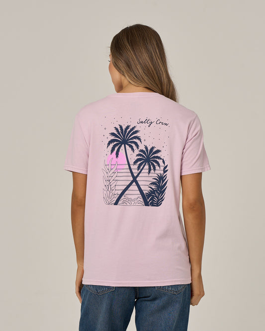 A woman with long hair, dressed in blue jeans and facing away from the camera, showcases the relaxed fit of the Salty Crew Moon Wake Boyfriend Tee in Pale Orchid—a light pink, short sleeve cotton jersey T-shirt featuring a graphic of palm trees, plants, and a sunset on the back along with the Salty Crew logo.