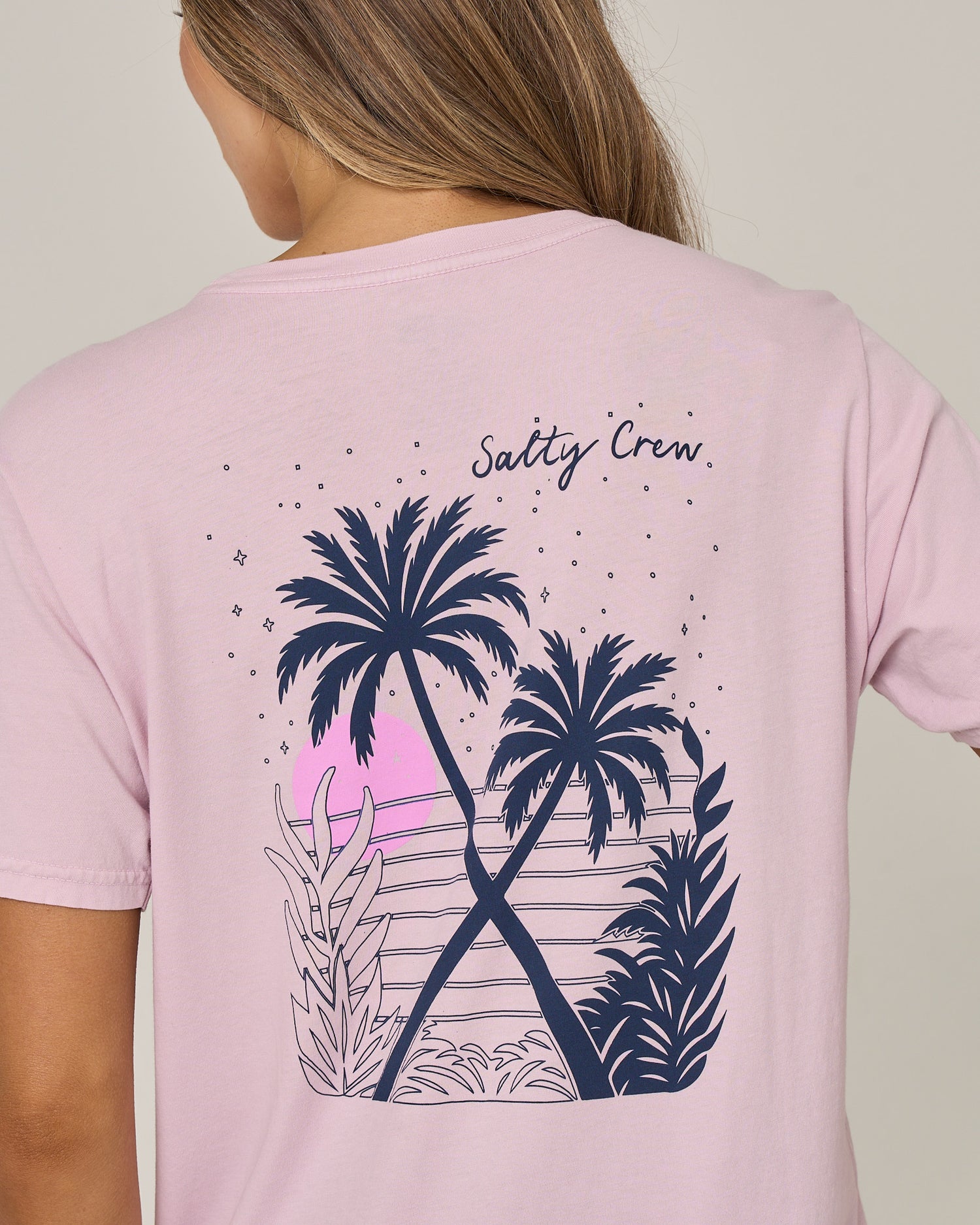 A person wears the Salty Crew Moon Wake Boyfriend Tee in Pale Orchid, a light pink, short sleeve cotton jersey T-shirt featuring an illustration of palm trees, plants, and a pink sunset on the back. “Salty Crew” is printed above the graphic, making this relaxed fit tee ideal for casual days.
