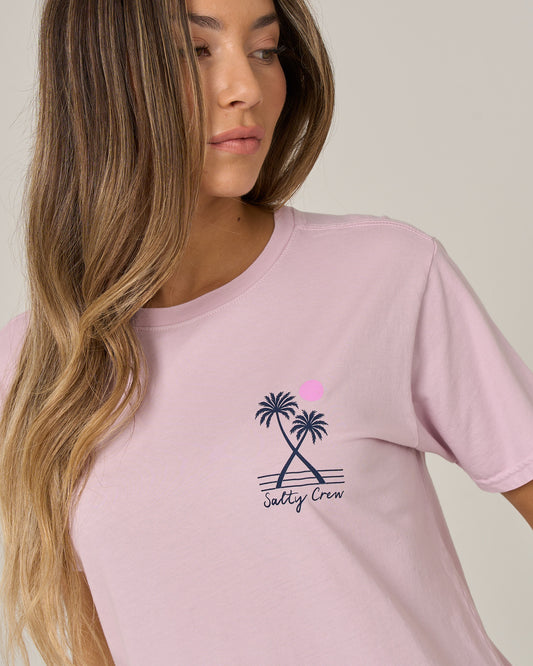 Woman with long, wavy hair looking to the side, wearing the Salty Crew Moon Wake Boyfriend Tee in Pale Orchid—a light pink short sleeve t-shirt featuring two palm trees and a pink sun graphic on the chest.
