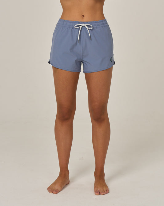 A person stands barefoot on a white background, wearing Salty Crew’s Beacons Short - Flint—a pair of light blue athletic shorts with a scallop hem and white drawstring. Made from recycled performance fibers and featuring quick dry fabric, only the lower torso and legs are visible.