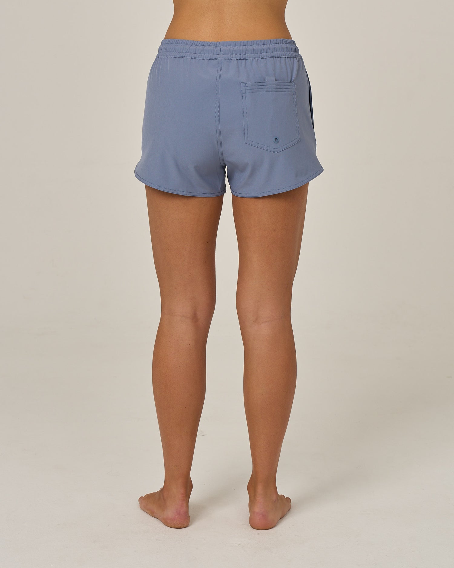 A person stands barefoot on a light surface, shown from behind, wearing the Salty Crew Beacons Short - Flint, which features a scallop hem and back pocket crafted from recycled performance fibers. The background is plain and neutral.
