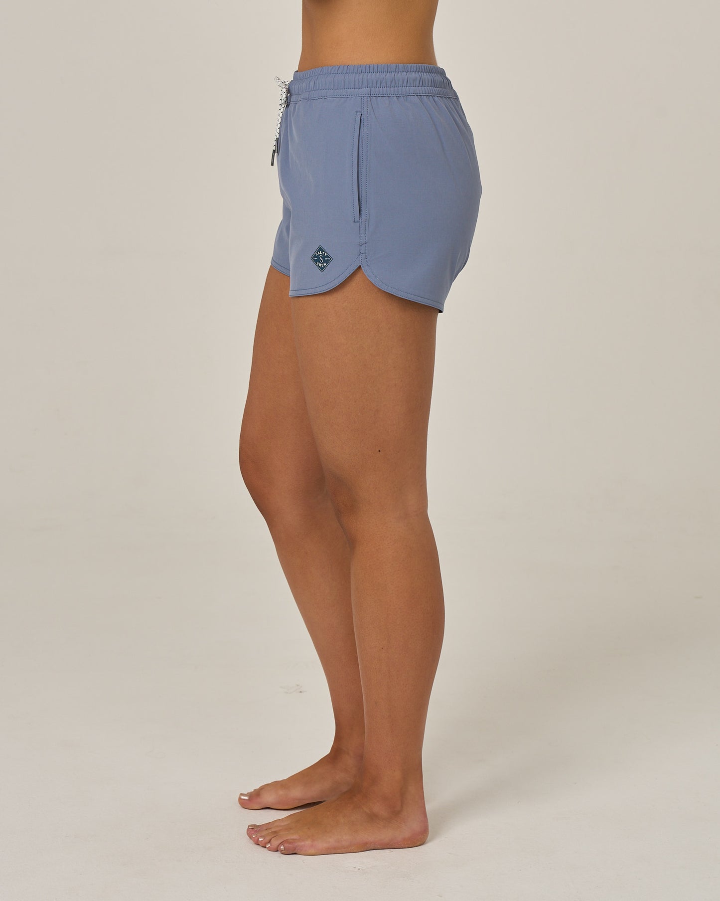 A person stands barefoot on a white background, wearing the Salty Crew Beacons Short - Flint, featuring an elastic waistband and a zippered pocket. Crafted from quick-dry fabric for comfort and performance, these light blue shorts showcase a scallop hem. Only the lower body is visible from the waist down.