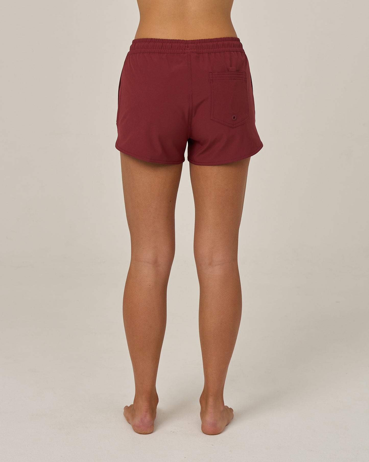 A person stands barefoot facing away, wearing the Salty Crew Beacons Short in maroon, featuring a scallop hem and back pocket, crafted from REPREVE recycled performance fibers, against a plain light background.