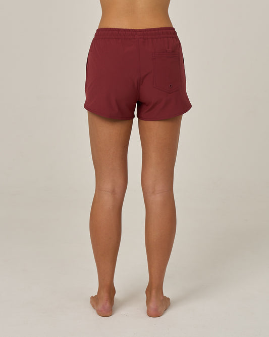 A person stands barefoot facing away, wearing the Salty Crew Beacons Short in maroon, featuring a scallop hem and back pocket, crafted from REPREVE recycled performance fibers, against a plain light background.