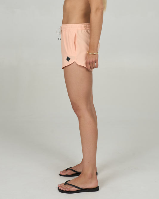 A person stands sideways against a plain light background, wearing Salty Crew's Beacons Short - Melon and black flip-flops. The shorts feature a scallop hem and are made with REPREVE recycled fibers. Upper body mostly out of frame.