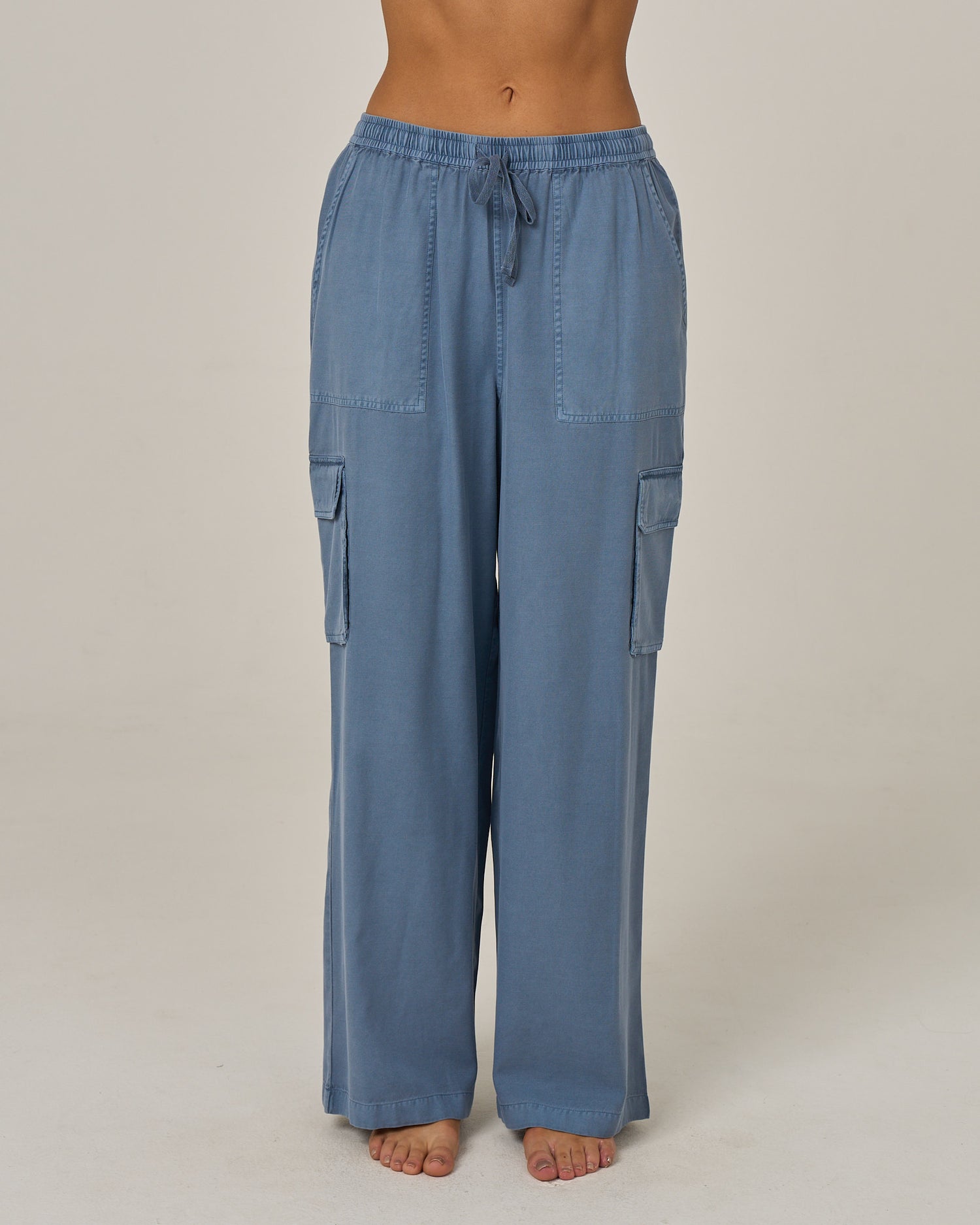 A person wearing the Salty Crew Driftwood Pant - Flint, a loose-fitting, light blue vintage cargo pant featuring side cargo pockets and an elastic waist. The full-length pants are shown on a barefoot model from mid-torso down against a plain background.