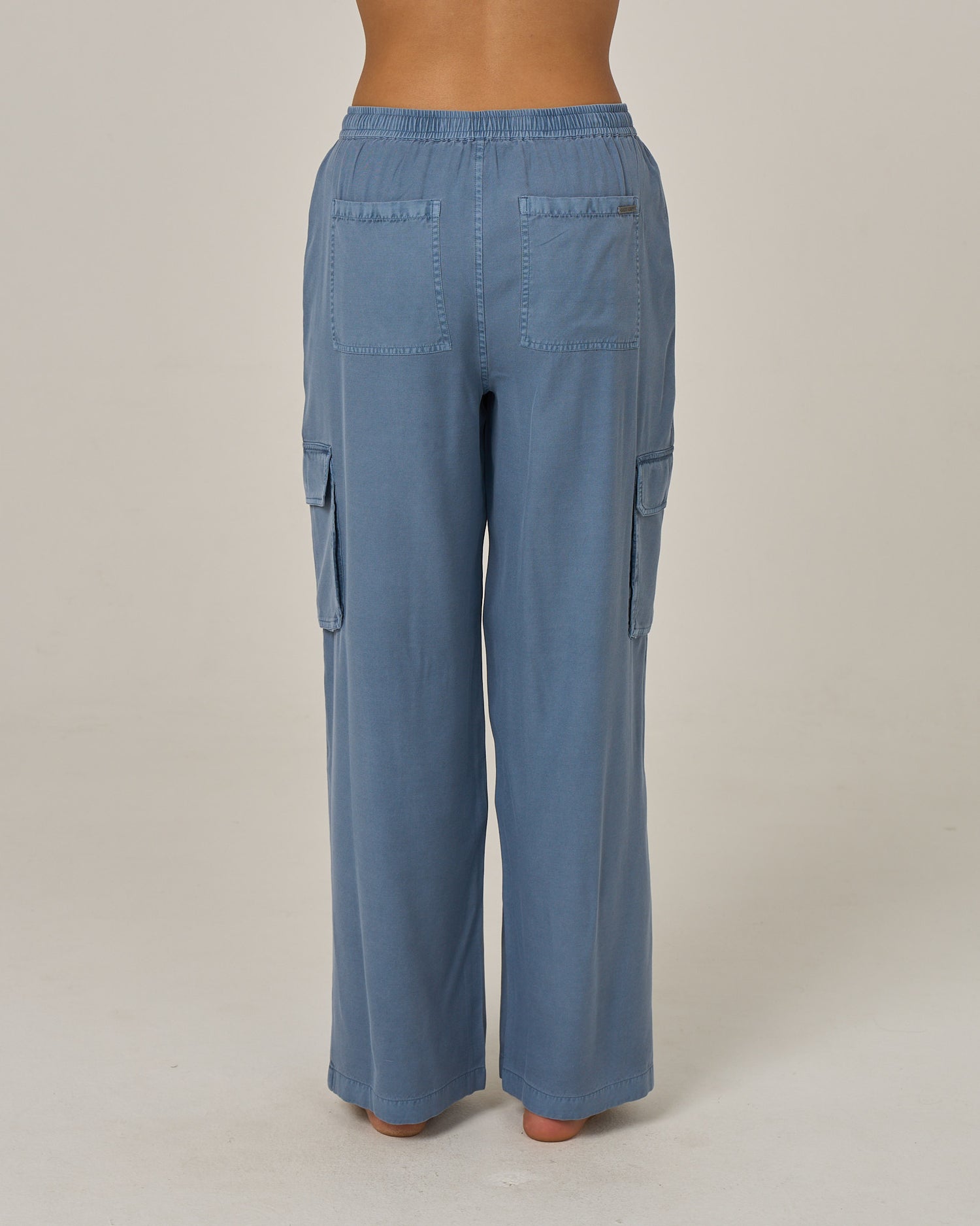 A person is shown from the back, standing barefoot against a plain light background, wearing the Salty Crew Driftwood Pant - Flint: a loose-fitting, light blue vintage cargo pant featuring an elastic waist and side cargo pockets.