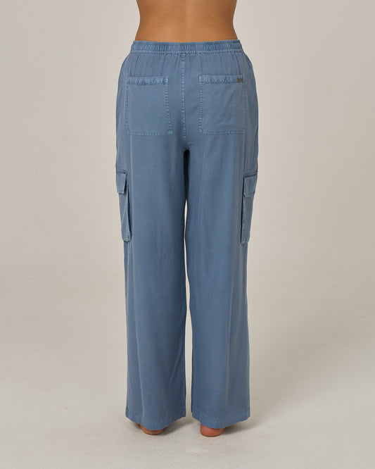 A person is shown from the back, standing barefoot against a plain light background, wearing the Salty Crew Driftwood Pant - Flint: a loose-fitting, light blue vintage cargo pant featuring an elastic waist and side cargo pockets.