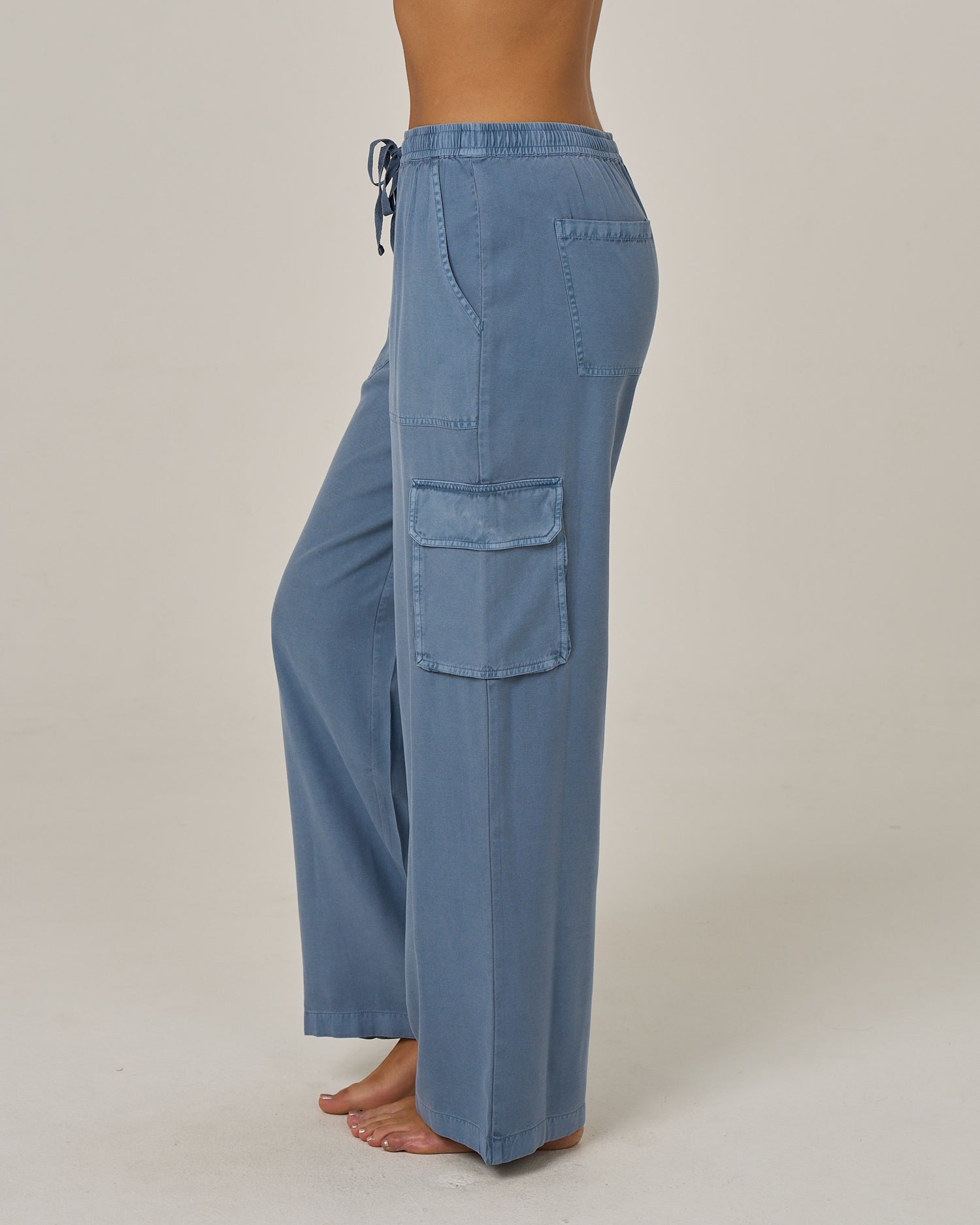 A person stands barefoot on a plain background, shown from the side, wearing the Salty Crew Driftwood Pant - Flint: loose-fitting vintage cargo pants in blue with an elastic waist and large side cargo pockets.