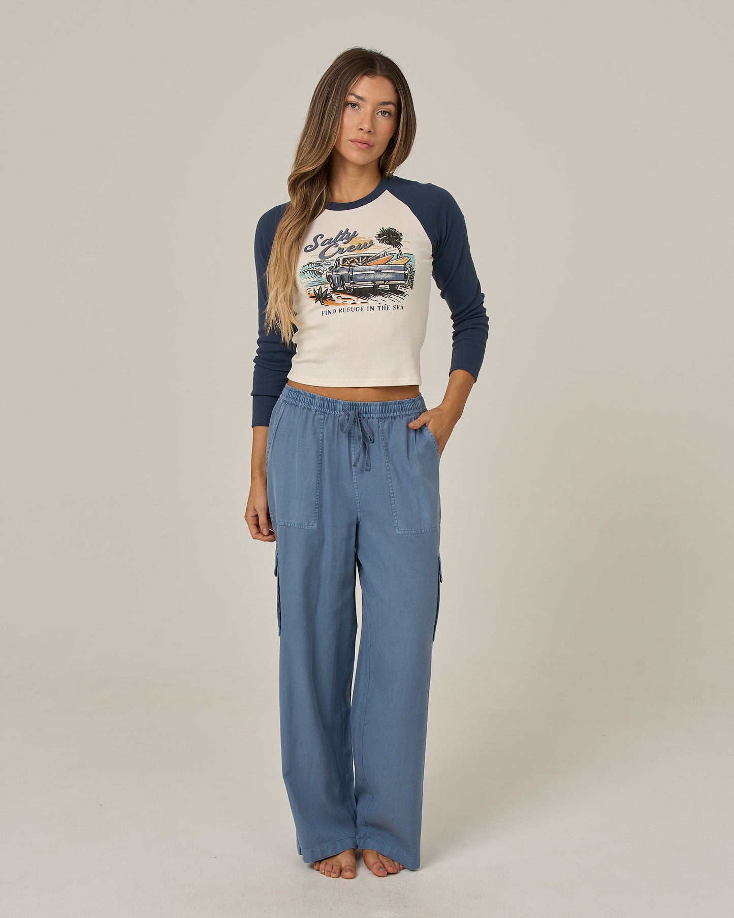 A woman stands barefoot in a neutral studio setting, wearing a graphic raglan shirt with blue sleeves and the Salty Crew Driftwood Pant - Flint, featuring an elastic waist and side cargo pockets.