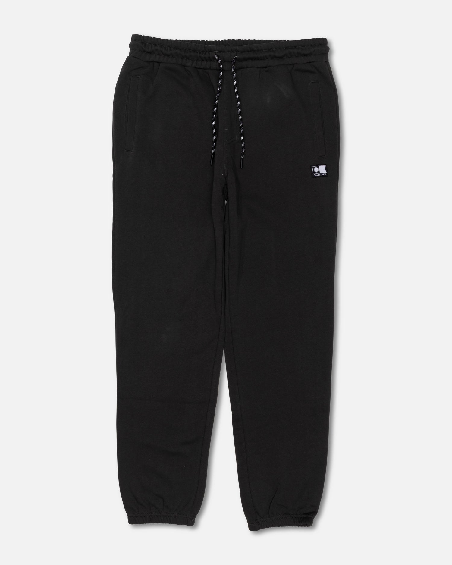 The Salty Crew Crewser Fleece Pant in Pirate Black features an elastic waistband with a drawstring, side pockets, and elastic cuffs at the ankles. These sweatpants are detailed with a small rectangular patch on the left side and subtle screenprinted artwork. Shown laid flat on a white background.